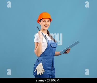 Smiling young handywoman in coveralls and hardhat making list of construction materials Stock Photo