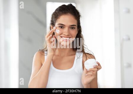 Attractive Young Woman Applying Face Cream Standing In Bathroom Indoor Stock Photo