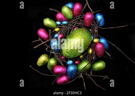 Foil wrapped multi coloured easter eggs in pink, green, blue and yelow in a natural nest made of wood, sticks and twigs, against a black background. Stock Photo