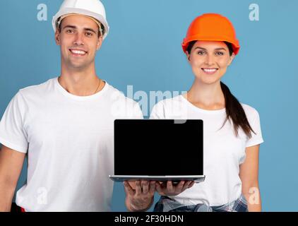 Happy Young Man And Woman In Hardhats Holding Laptop With Black Screen Stock Photo