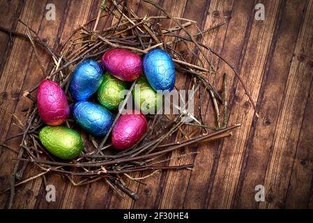 Foil wrapped colourful easter eggs in pink, green, blue and yelow in a natural nest made of sticks and twigs, against a multi grain brown wooden backg Stock Photo
