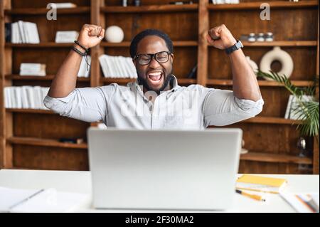 Excited lucky African American student or office manager sitting with laptop at the desk, raised fists up celebrating success, screaming yes, passed test with high score, got promoted, won an award Stock Photo