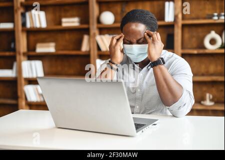 Stressed young African-American entrepreneur in protective face mask, touching massaging temples, having headache feeling tired from constant work on laptop while sitting in office with bookshelves Stock Photo