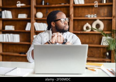 Serious thoughtful young African American man with dreadlocks sitting at desk in the office library and looking away, taking break from working on laptop, pondering project plan, thinking of new ideas Stock Photo