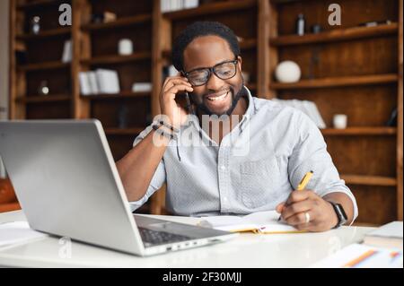 Successful African-American businessman in glasses working in the office, sitting at the desk with laptop, holding smartphone, talking with client. Friendly manager company member making phone calls Stock Photo