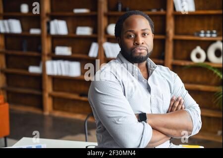 Portrait of a serious young confident African American male entrepreneur standing with arms folded in the library or and looking at the camera, intelligent black IT guy posing with arms crossed Stock Photo