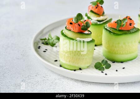 Cucumbers rolls with soft cheese, pieces of salted salmon, microgreens and black sesame served on a white plate. Holiday vegetable appetizers. Selecti Stock Photo