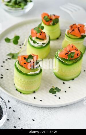 Cucumbers rolls with soft cheese, pieces of salted salmon, microgreens and black sesame served on a white plate. Holiday vegetable appetizers. Selecti Stock Photo