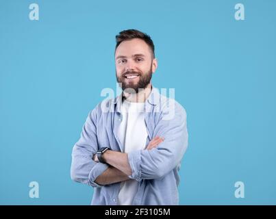 Portrait of positive young guy posing with crossed arms and smiling at camera on blue studio background Stock Photo