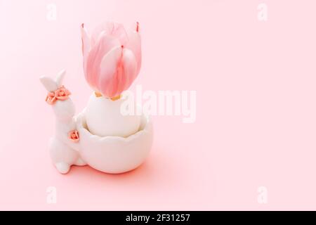 Easter egg in stand with ceramic easter bunny figurine and spring tulip flower on pastel pink background Stock Photo