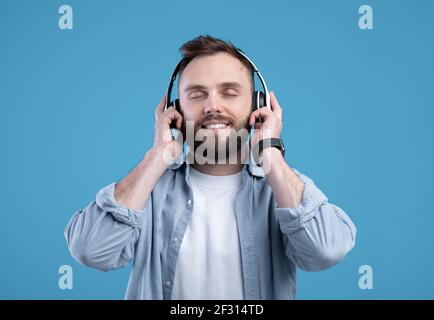 Attractive bearded man in casual outfit listening to music, wearing headphones on blue studio background Stock Photo