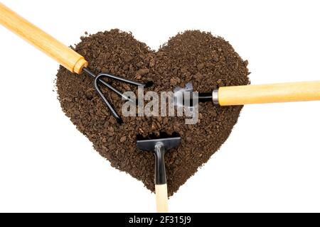 Various garden hand tools in a heart-shaped pile of soil isolated on white background. Gardening obsession concept. Stock Photo