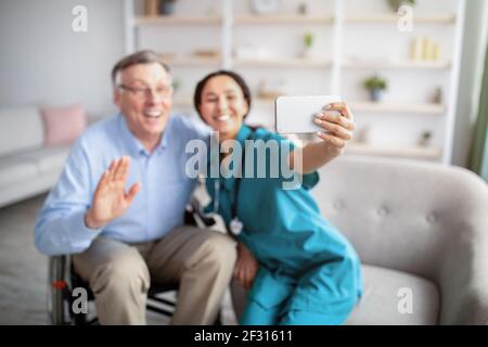Cheerful disabled senior man in wheelchair and his nurse taking selfie indoors, selective focus Stock Photo