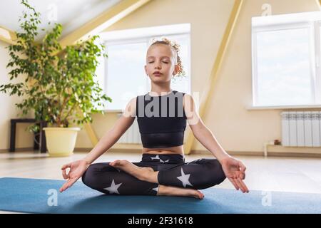A little girl in sportswear, practicing yoga, is engaged in meditation sitting in the lotus position in the room. Child yoga concept. Stock Photo