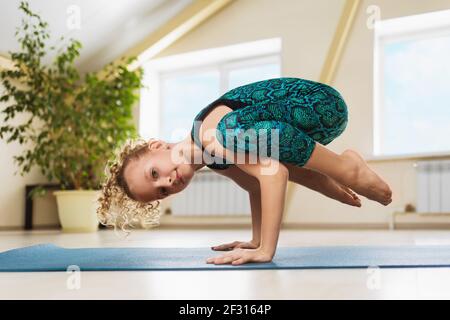 Little beautiful blonde girl practicing yoga in the studio, doing handstand exercise, kakasana or crow pose on a gymnastic rug. Stock Photo