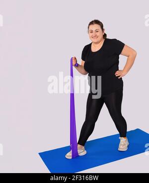 Smiling athletic large woman training body at home, active sports workout. Fat female athlete in activewear doing forward lunges exercise with elastic Stock Photo