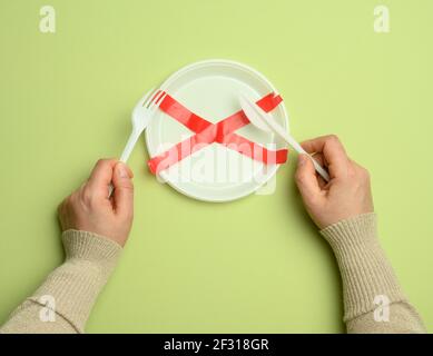 Empty white plastic plate sealed with red tape and two hands holding a knife and fork, green background Stock Photo