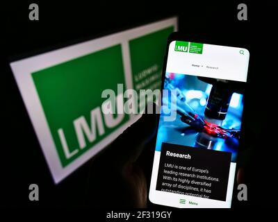Person holding cellphone with web page of German education institution LMU Munich on screen in front of logo. Focus on phone display. Unmodified photo. Stock Photo