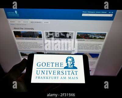 Person holding cellphone with logo of German education institution Goethe University Frankfurt on screen in front of website. Focus on phone display. Stock Photo