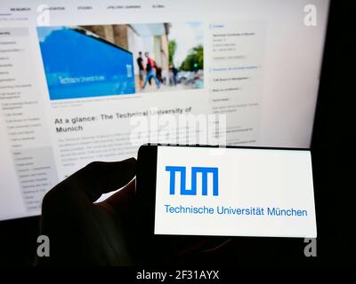 Person holding smartphone with logo of education institution Technical University of Munich on screen in front of web page. Focus on phone display. Stock Photo