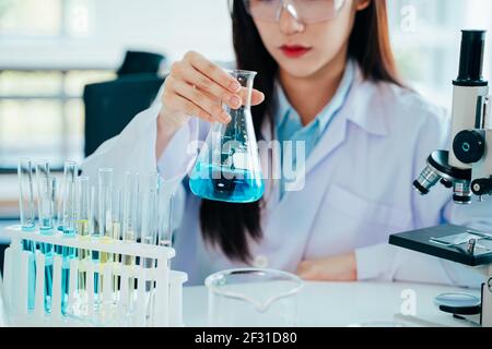 Focused young and beautiful female Asian scientist wearing labcoat and safety eyewear doing medical research in lab with test tubes and microscope Stock Photo
