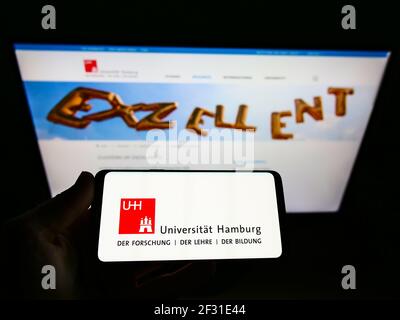 Person holding smartphone with logo of German education institution University of Hamburg on screen in front of website. Focus on cellphone display. Stock Photo