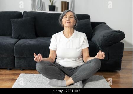 A peaceful and calm senior female practicing yoga at home, an older woman sits in lotus position, a middle aged lady meditates. Health and wellbeing concept Stock Photo