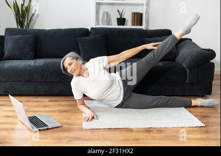 Active and healthy retiree woman is exercising with training videos on the laptop, senior female stretching legs lying on the floor and watching sports video tutorial, takes part in the marathon Stock Photo