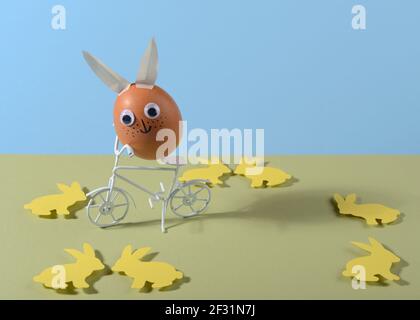 Happy Easter concept. Easter egg with eyes and rabbit ears on bicycle. Copy space. Paper cut. Stock Photo