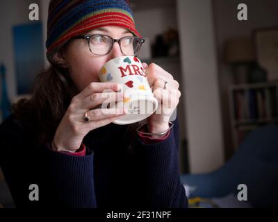 A middle-aged woman wearing a knitted hat drinks from a mug with decorated with hearts and 'Mrs" on the side. Stock Photo