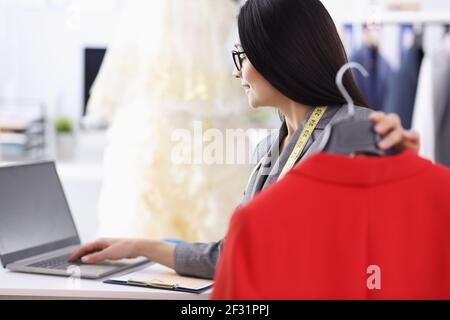Woman dressmaker is holding a red jacket and working on laptop Stock Photo