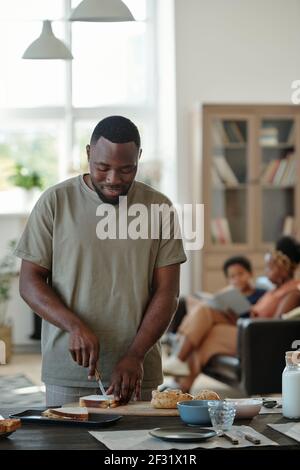 Young African man in casualwear preparing sandwiches for breakfast while standing by table in the kitchen against his wife and son reading book Stock Photo