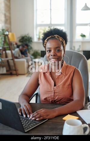 Successful young African female freelancer looking at you during work in front of laptop while sitting in armchair by table against her family Stock Photo