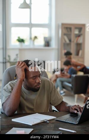 Pensive young African man with pencil in hand sitting by table in front of laptop and scrolling through online data against his family Stock Photo