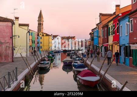 The Venetian Island of Burano, boats moored on a canal, colorful houses and shops at dusk with people on the sidewalks. Stock Photo