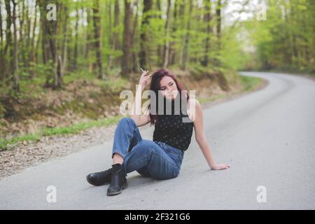 A young female with colored hair smoking while sitting on the road near a forest Stock Photo