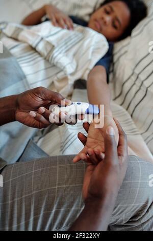 Hands of young man of African ethnicity holding thermometer while measuring body temperature of his sick little son lying in bed Stock Photo
