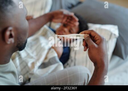 Hand of young careful man looking at thermometer after measuring body temperature of his sick little son and touching his forehead Stock Photo