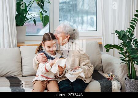 Loving granny sitting on a couch with her granddaughter. they are embroidering on a loop together. She's proud of her, kissing her hair. Stock Photo
