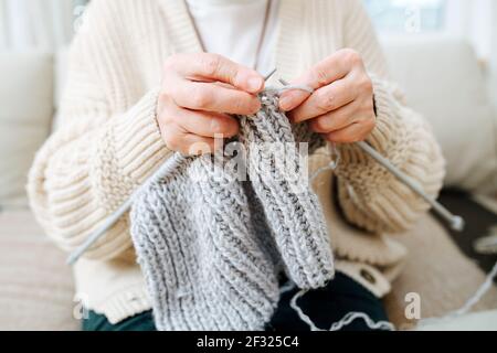 Granny sitting on a couch at home, knitting with needles, using grey wool. Cropped, no face. Stock Photo