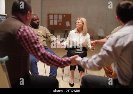 Several intercultural patients of mental support group holding by hands during session while sitting in circle and listening to one another Stock Photo