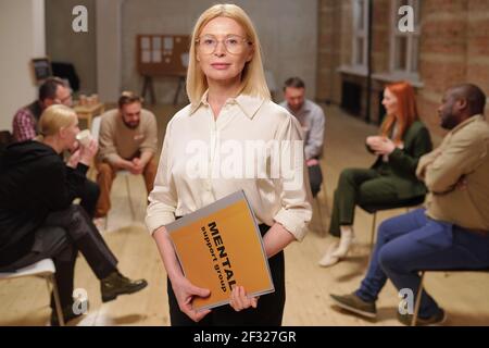 Confident mature female counsellor in eyeglasses and smart casualwear holding journal of mental support group while standing against patients Stock Photo