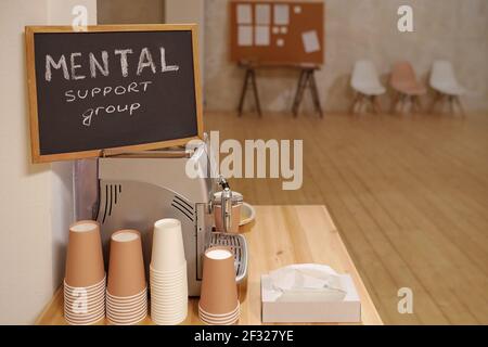Small blackboard with notice saying mental support group standing on coffee machine with several stacks of paper glasses near by Stock Photo