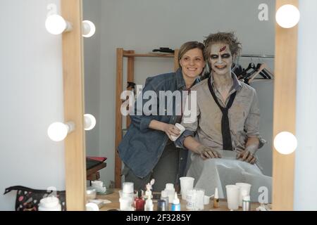 Reflection in mirror of female makeup artist and young grinning spooky man with zombie greasepaint on his face, neck and hands Stock Photo