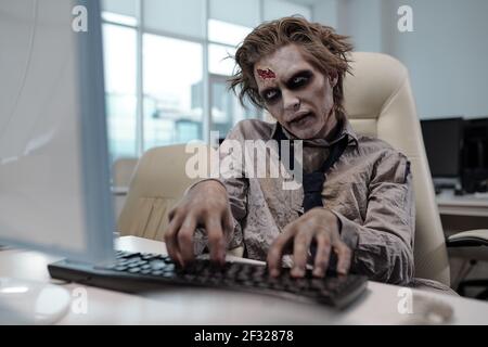 Spooky businessman with zombie body painting looking at computer screen and pressing keyboard buttons in office environment Stock Photo