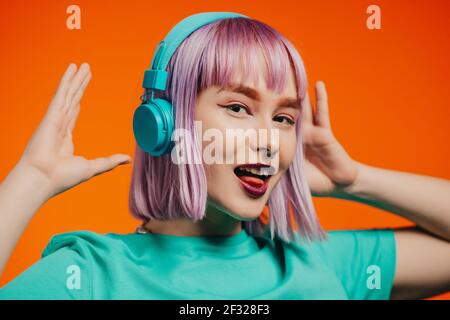 Exceptional woman with dyed violet hair listening music in headphones and singing on orange background. Unique hipster girl dancing Stock Photo