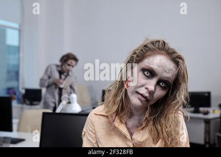 Young zombie businesswoman standing in front of camera on background of desks with computer monitors and dead male colleague Stock Photo