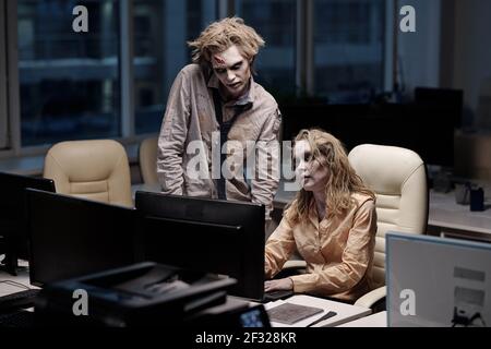 Young spooky zombie businessman standing next to female colleague sitting by desk in front of computer monitor during teamwork Stock Photo