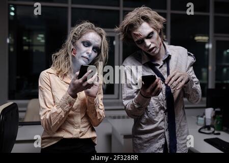 Dead and spooky businesspeople with zombie greasepaint on their faces and hands scrolling in smartphones in office in the midnight Stock Photo