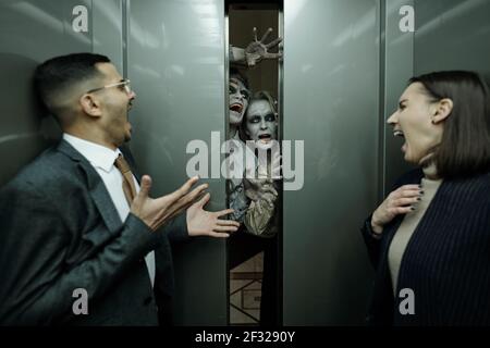 Young contemporary thrilled office managers in formalwear shouting in horror in moving elevator while two zombies trying to get inside Stock Photo